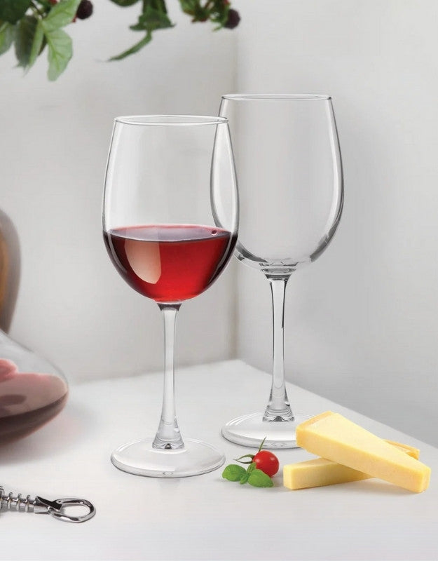 Two wine glasses with red wine and cheese on a white surface. Two elegant wine glasses, one filled with red wine and one empty, placed beside cheese slices and a cherry tomato on a white table.